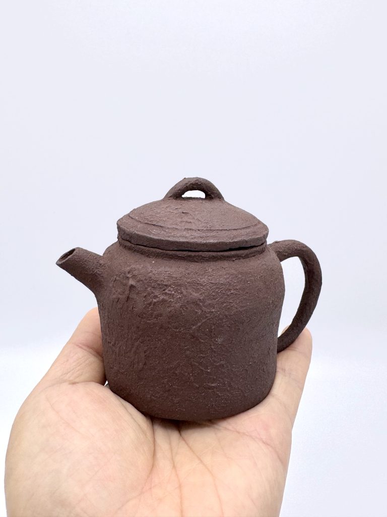 A small dark reddish brown zisha teapot held in an open hand, showing its scale. It sits roughly within the width of the palm. Spout on the left, round handle on the right. The clay surface is rough and unglazed.