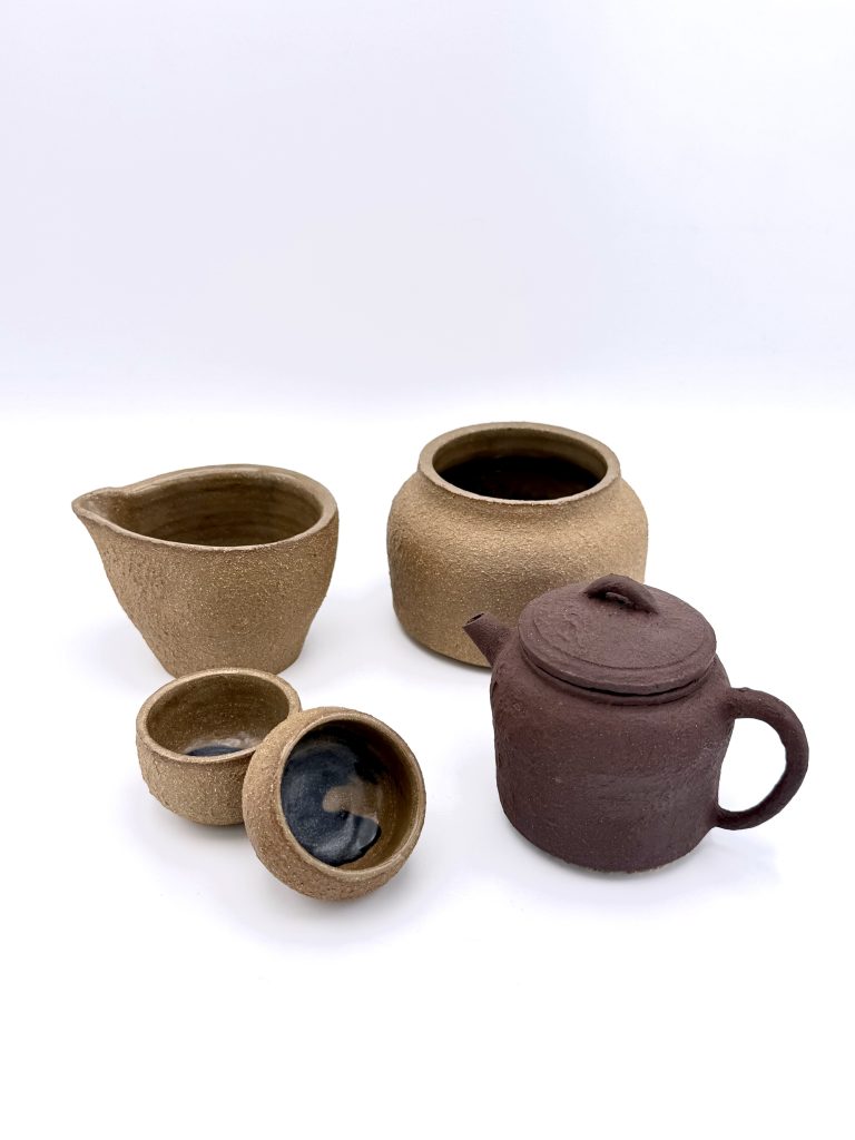 The full tea set on a white background. At back left, a pale tan pouring pitcher; to its right, the taller tan waste water jar. In front, two small tan tea cups on the left and the dark brown zisha teapot on the right. The cups have a dark glazed interior.